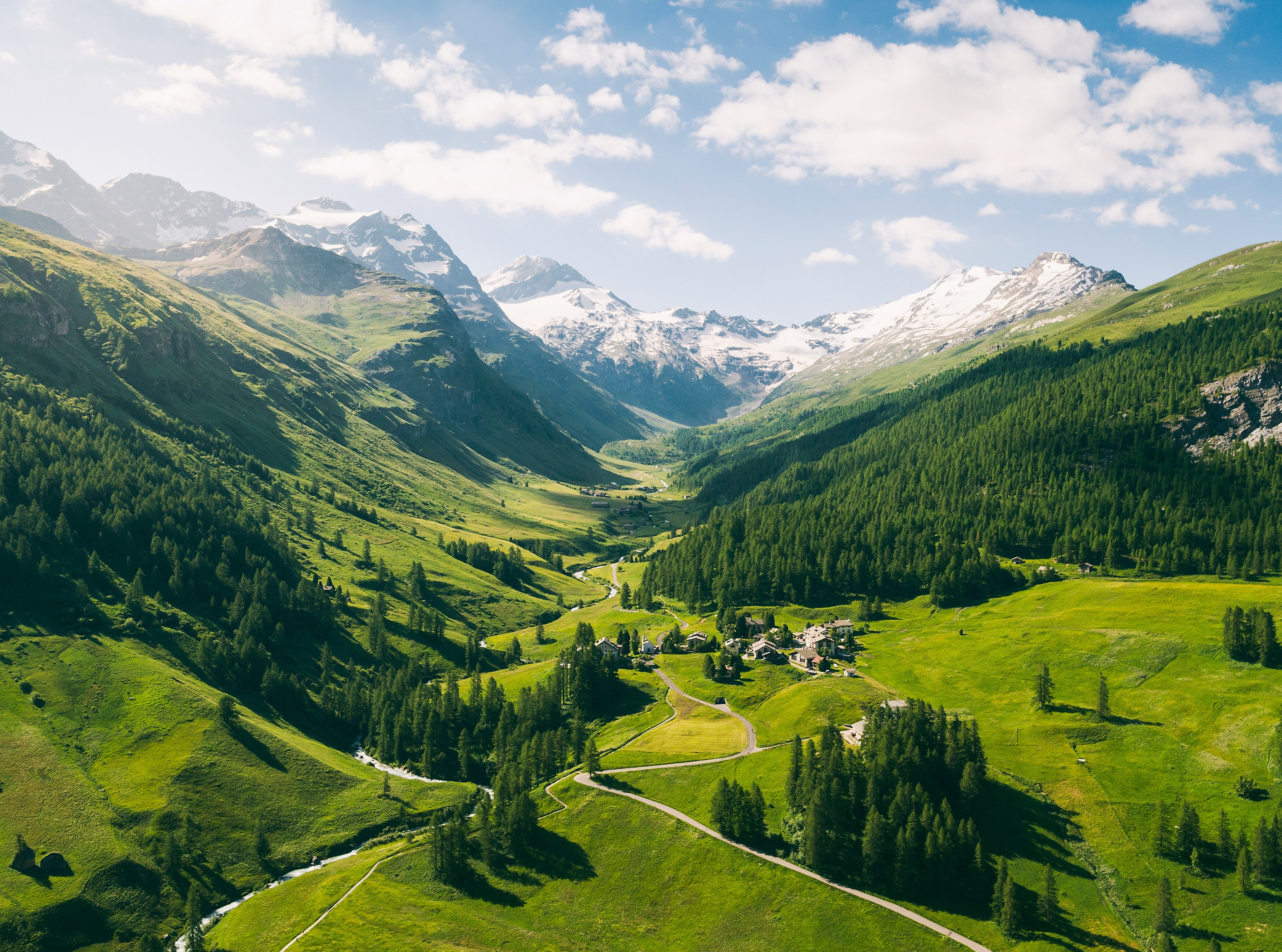 Aerial view of the Fex Valley in summer with lush green meadows, mountain forests, a small hamlet and snow-capped peaks in the background