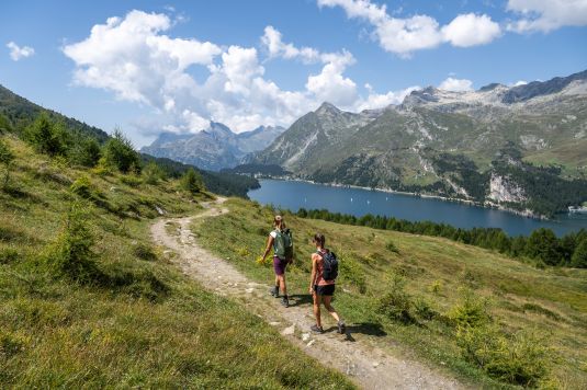 Zwei Wanderinnen auf einem Bergpfad oberhalb des Silsersees im Oberengadin mit Blick auf Berge und türkisblaues Wasser